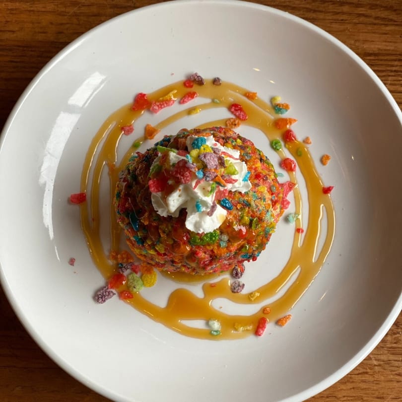 Colorful dessert ball covered in fruity cereal, topped with whipped cream and caramel drizzle.