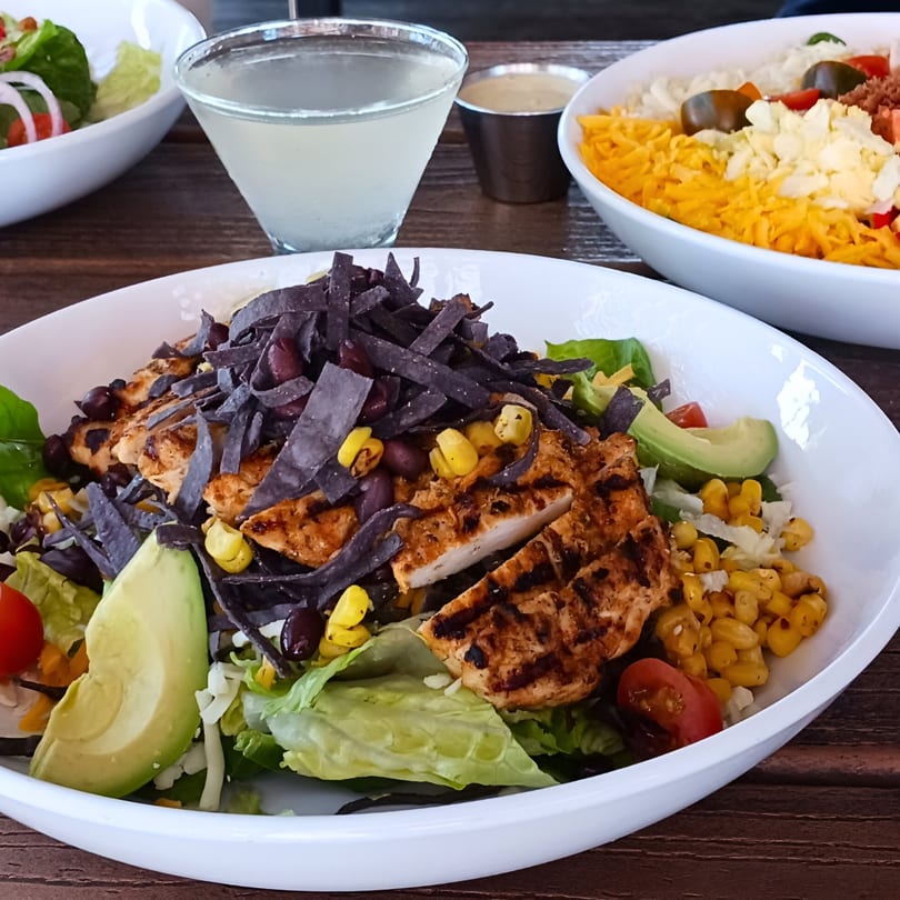Grilled chicken salad with avocado, corn, black beans, and tortilla strips, with a drink and another salad.